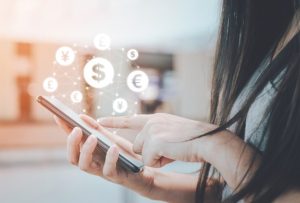 Mulher utilizando um smartphone com ícones de moedas e símbolos de diferentes moedas internacionais flutuando na tela, representando transações financeiras digitais. A imagem ilustra o conceito de empréstimo pessoal via carteira digital com praticidade e conectividade.
